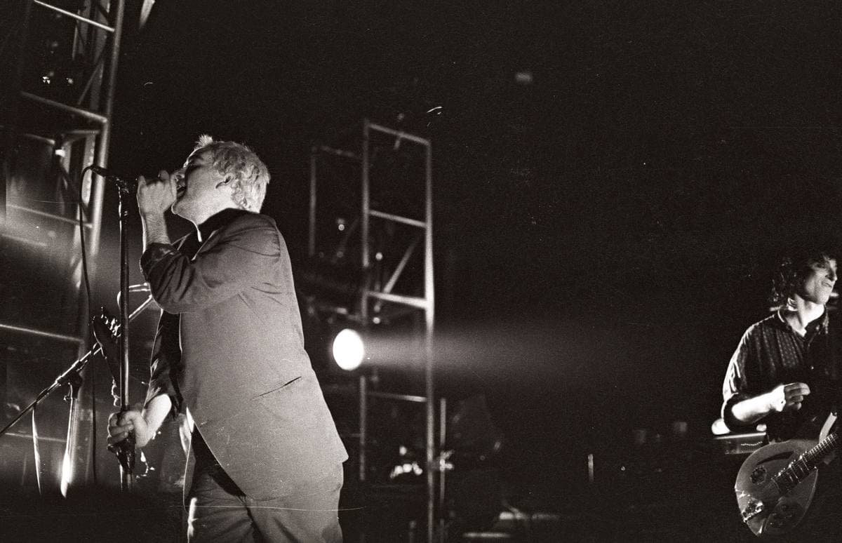 A singer under a single spotlight at a microphone stand, a guitarist at the right edge, black and white