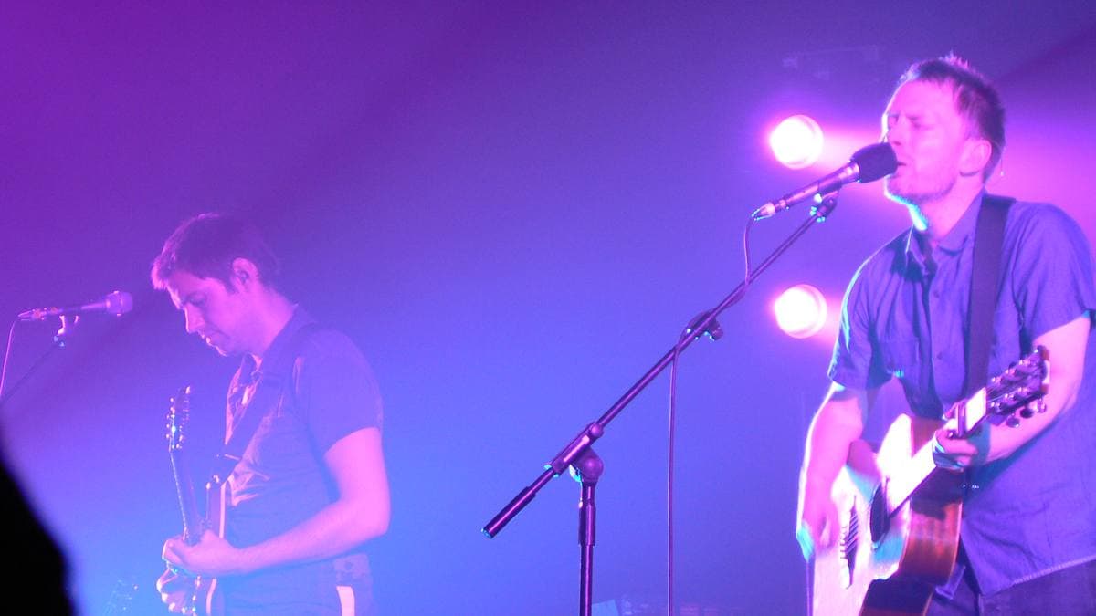 Ed O’Brien and Thom Yorke playing guitars at microphones under violet light