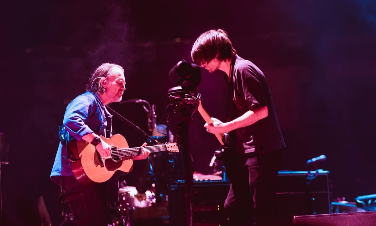 Thom Yorke with an acoustic guitar facing Jonny Greenwood on a dark stage lit pink