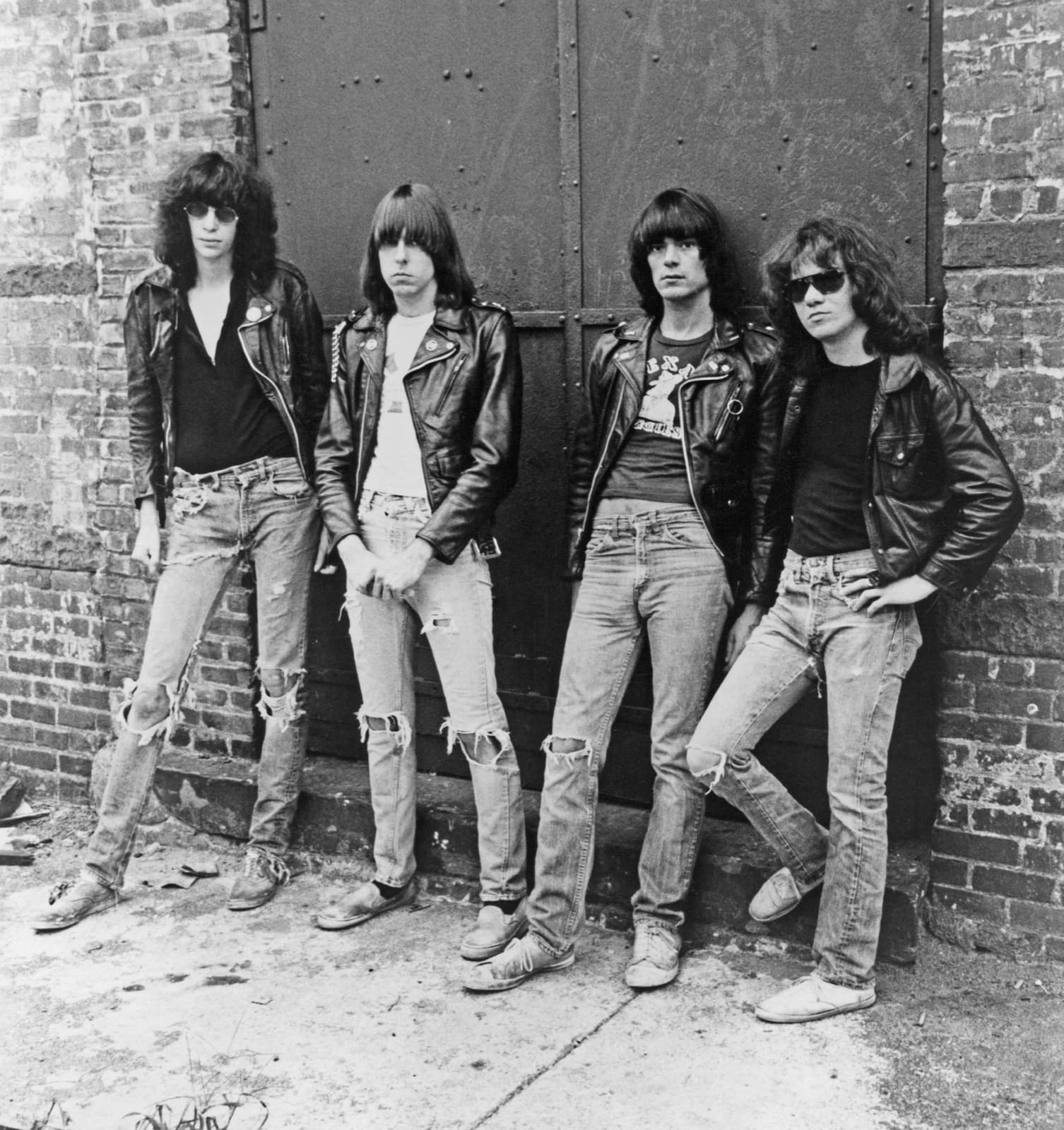The four Ramones in leather jackets and torn jeans, leaning against a steel door in a brick wall, in black and white