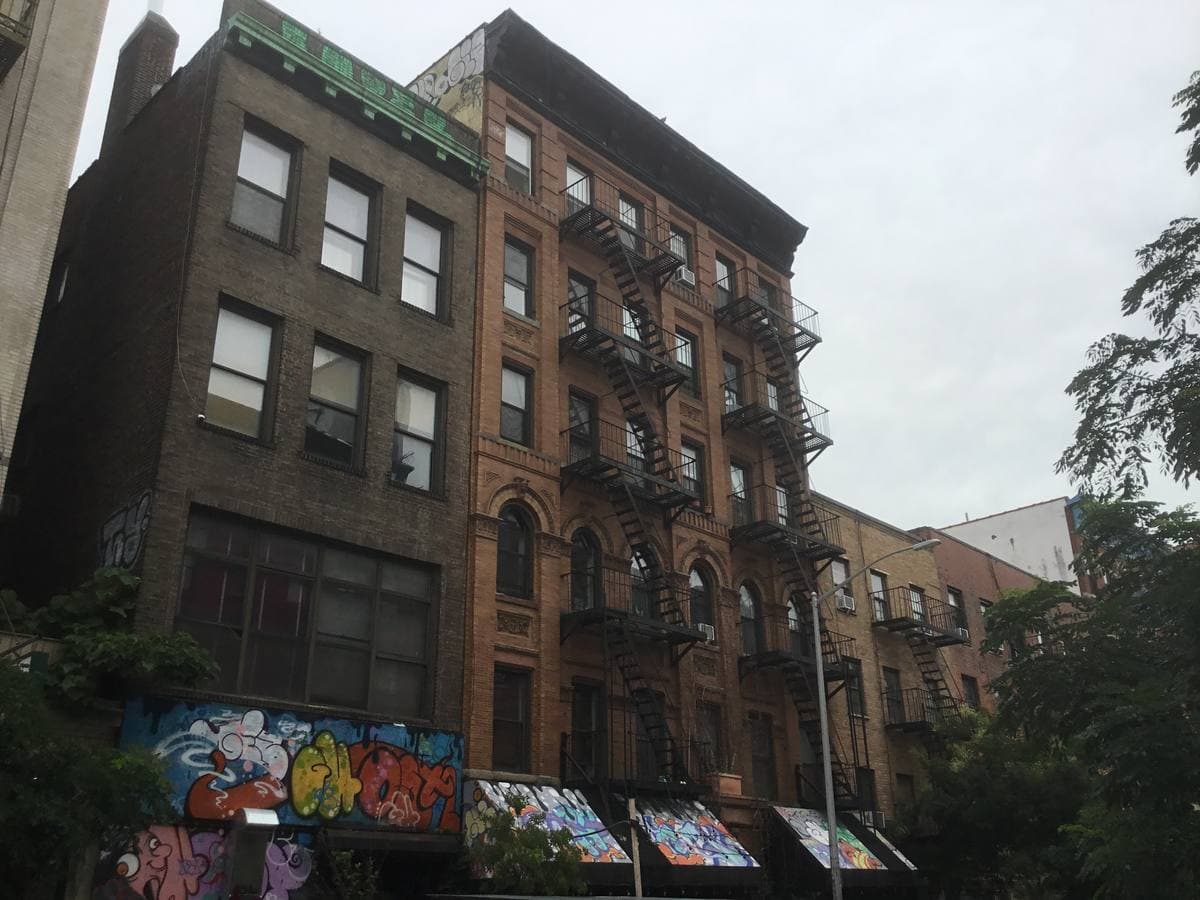 A tree-lined East Village street of brick tenements with fire escapes in summer