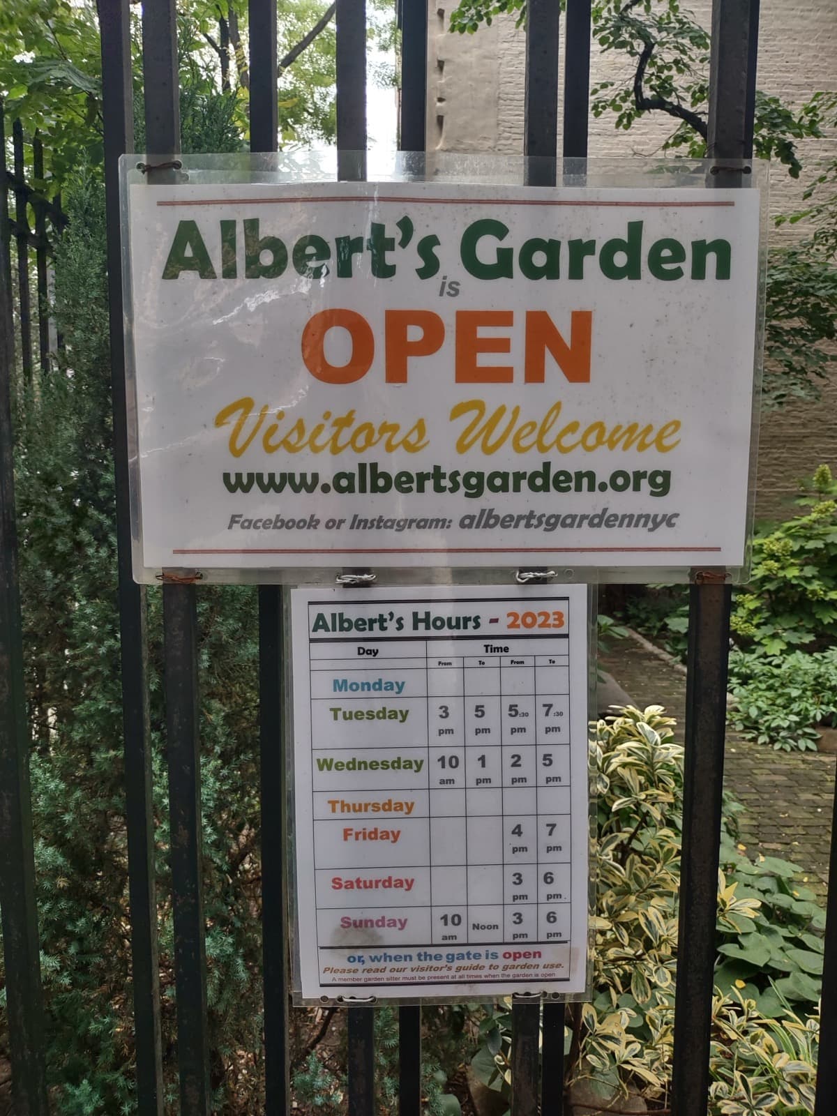 A sign reading Albert’s Garden is open, visitors welcome, on an iron fence in front of greenery