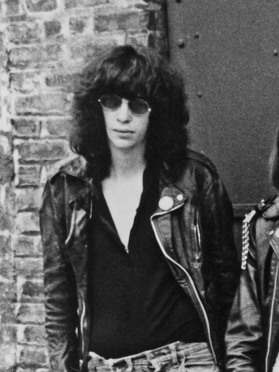 Joey Ramone, tall, in round dark glasses and a leather jacket, leaning on a brick wall