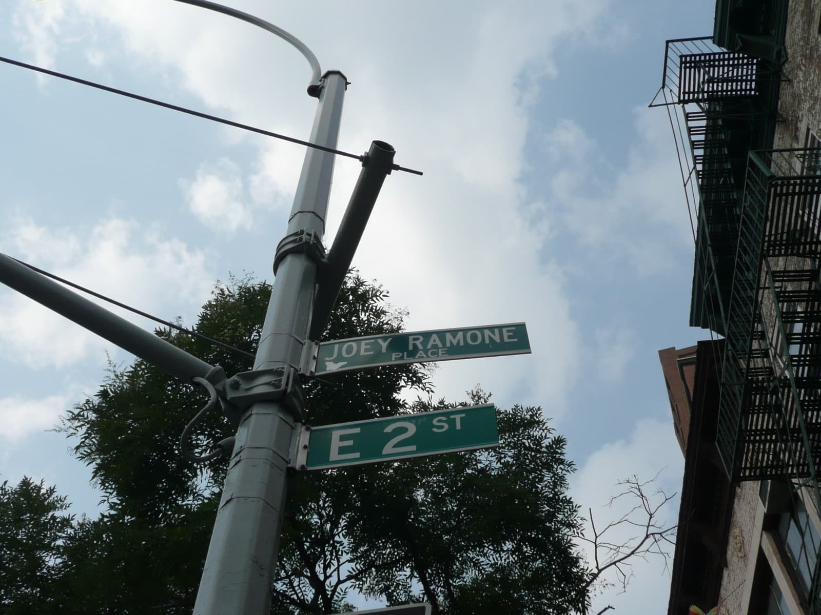 Green street signs for Joey Ramone Place and East 2nd Street on a lamppost against a blue sky