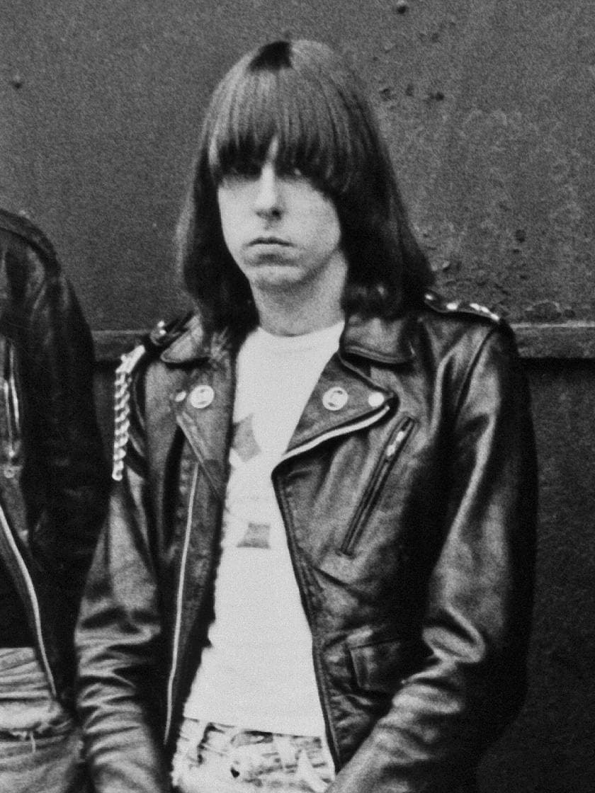 Johnny Ramone with a straight fringe, white T-shirt and leather jacket against a steel door