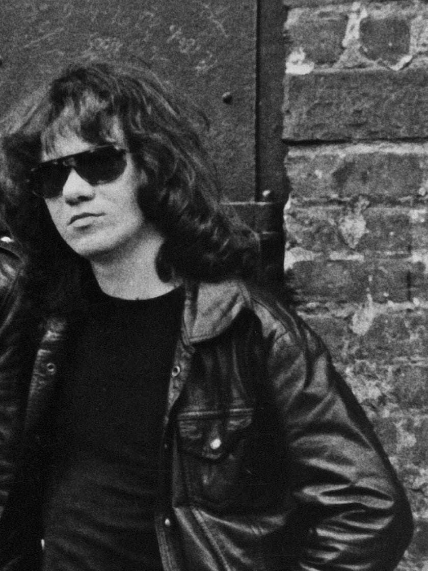 Tommy Ramone in dark glasses and a leather jacket against a brick wall