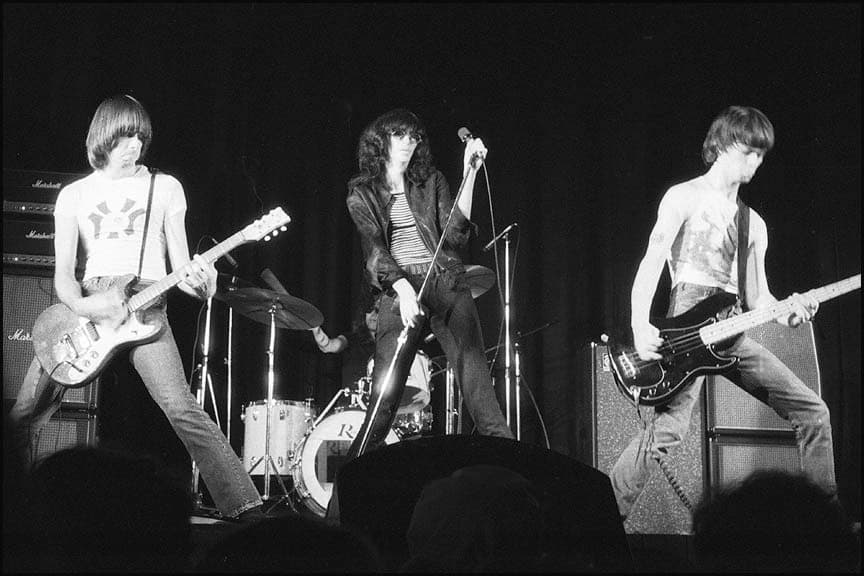 Johnny, Joey and Dee Dee Ramone on a dark stage, with Tommy at the drums behind them, in black and white