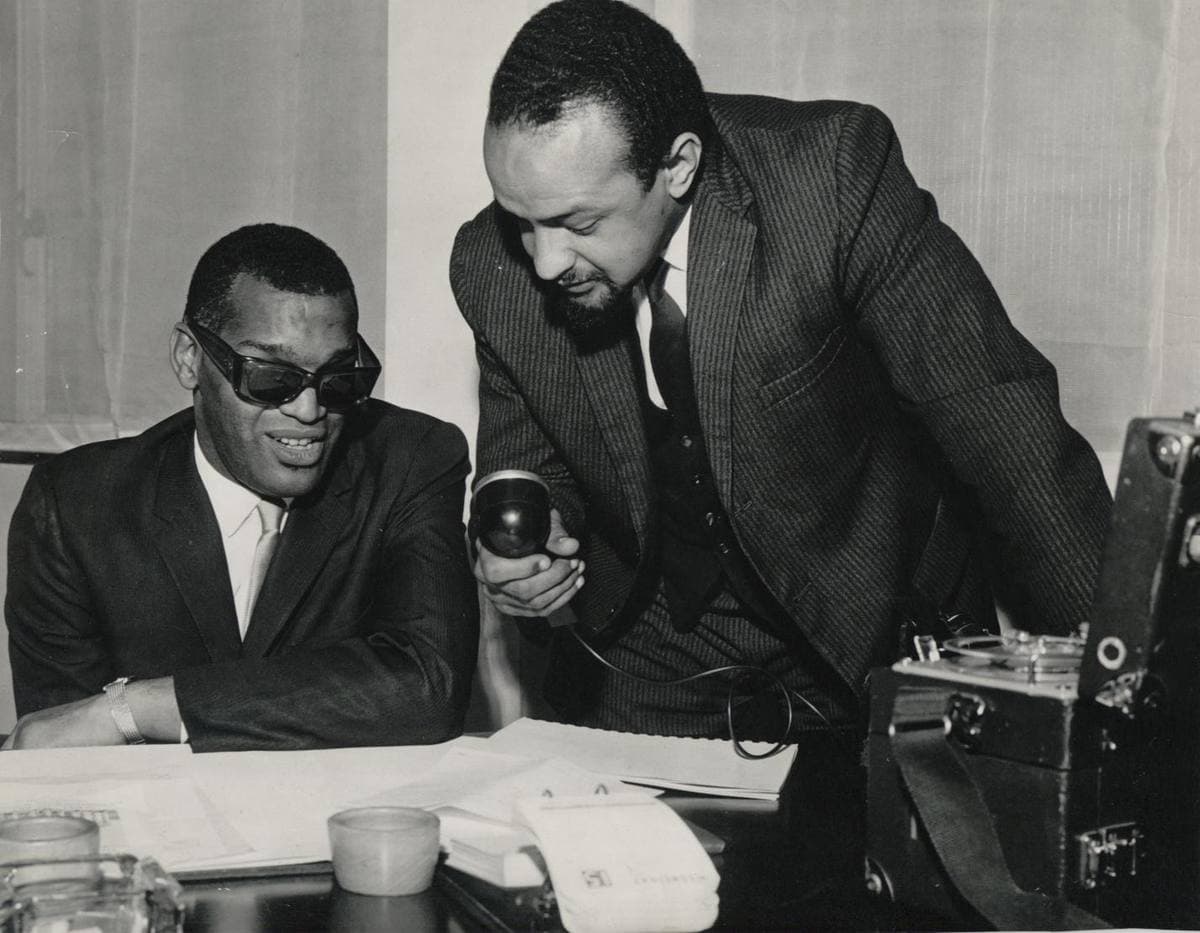 Ray Charles in dark glasses smiling at a desk while Andrew Salkey leans over him with a microphone, a tape recorder beside them