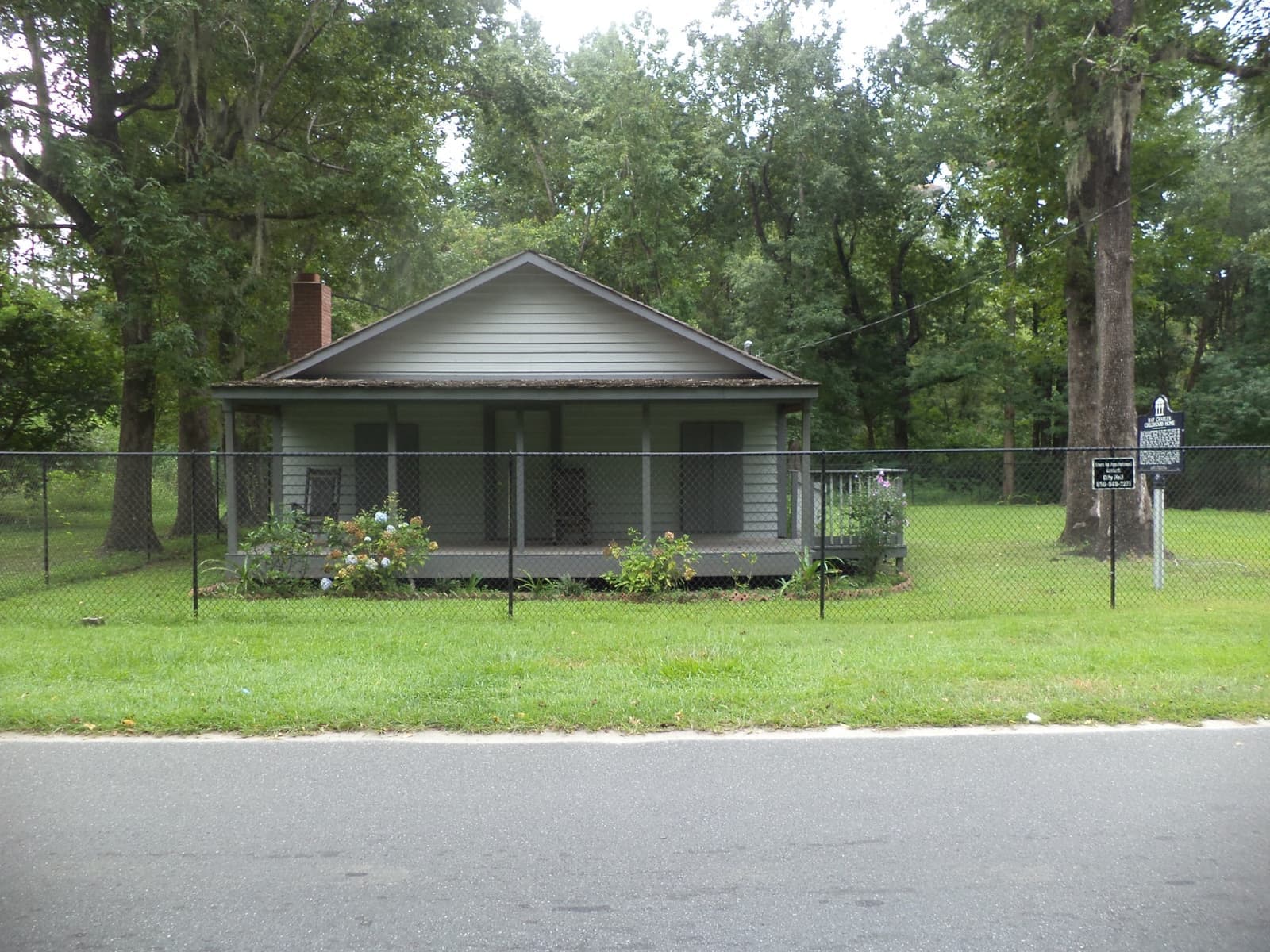 A small grey wooden house with a front porch behind a low fence, under tall trees