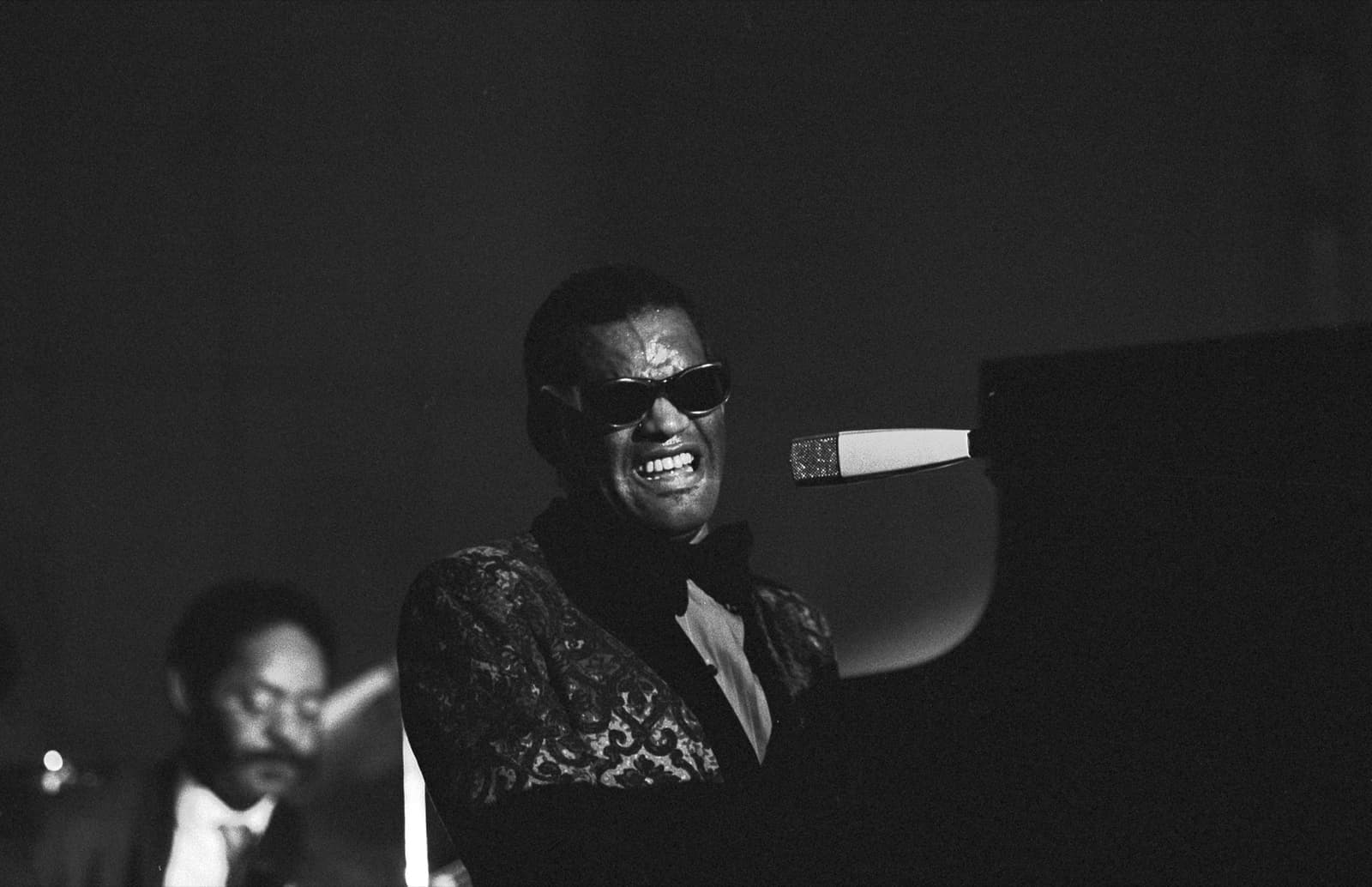 Ray Charles singing at a grand piano on a dark stage, head tilted back, in a patterned jacket