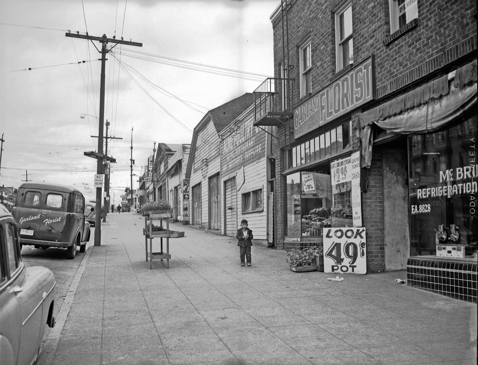 Black-and-white street of shopfronts and awnings, a parked car and a boy on the pavement