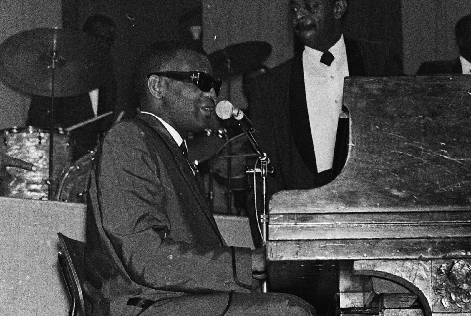 Ray Charles at a grand piano singing into a microphone, a band member in a dinner jacket behind him
