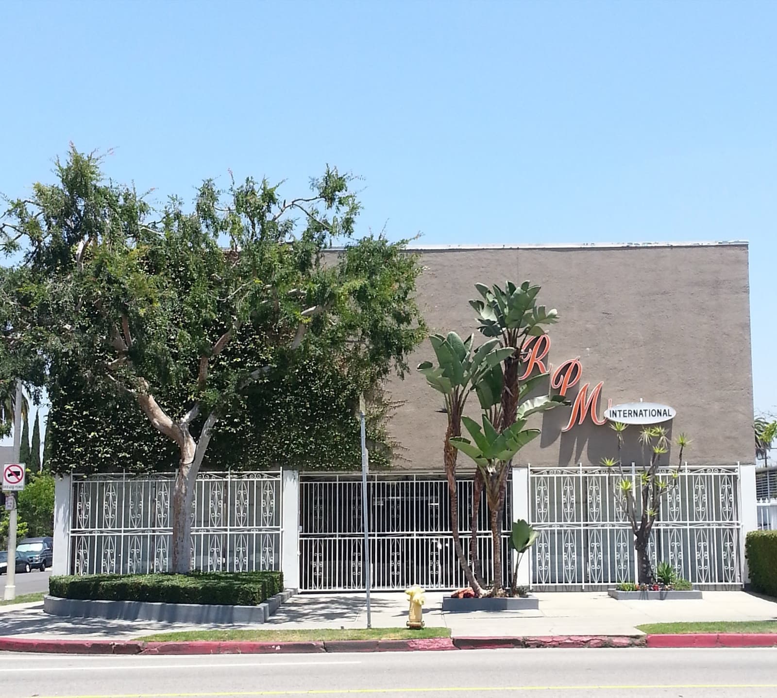 A low building with a decorative white grille fence, palms and a script sign on a blank wall