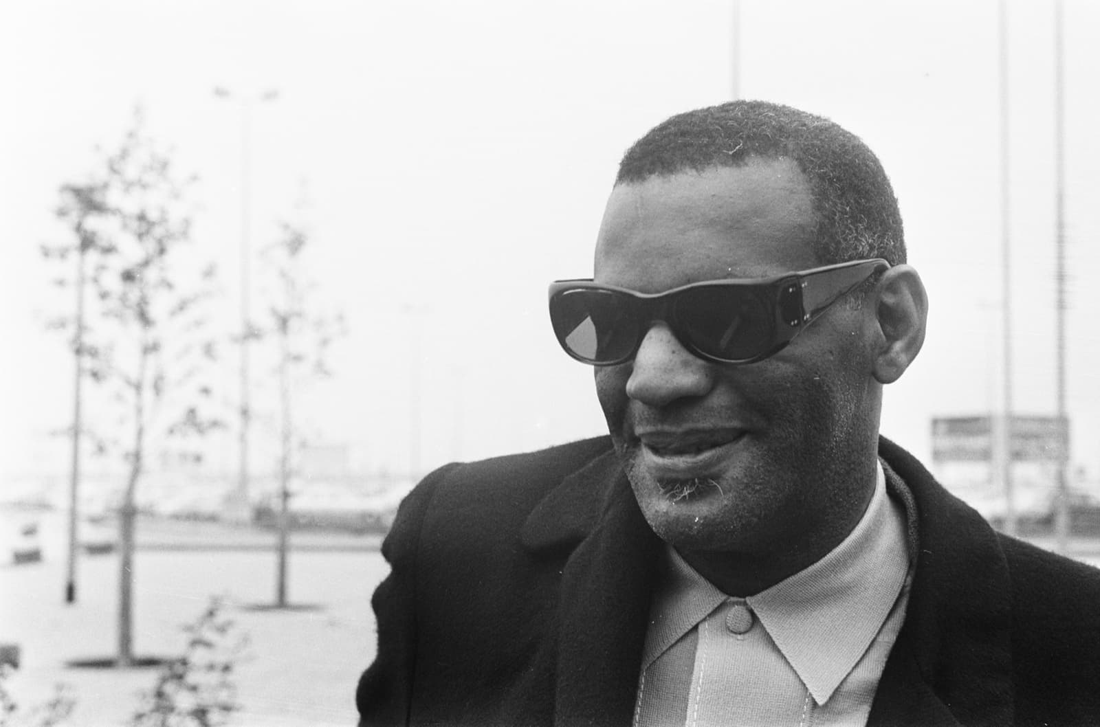 Ray Charles in dark glasses and an overcoat, smiling, outdoors at an airport