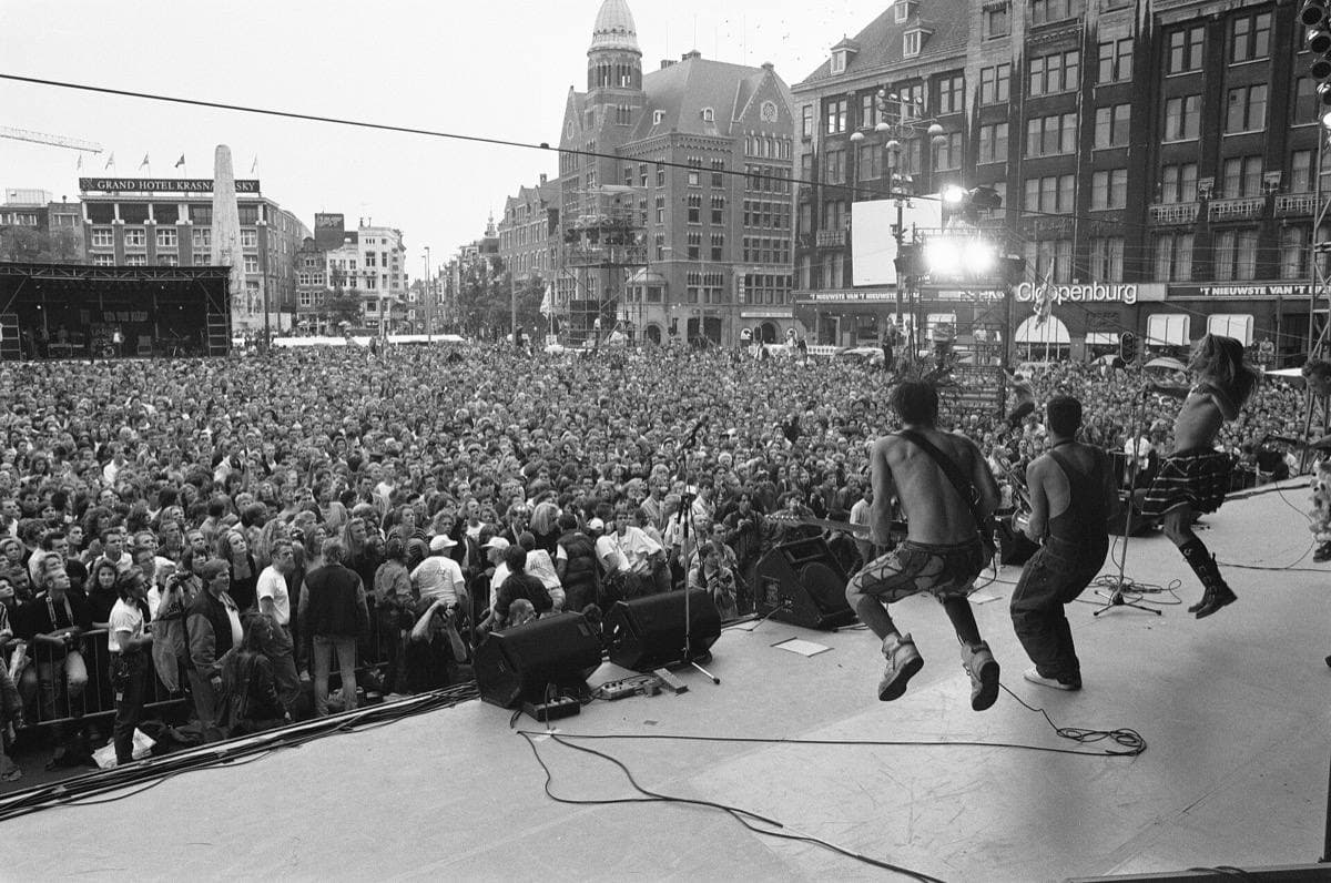 A crowd filling Dam square seen from the back of a stage, with musicians mid-jump in the foreground