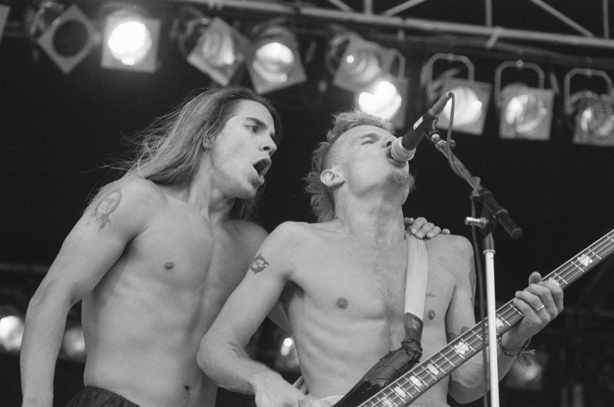 Anthony Kiedis and Flea, shirtless, singing into one microphone under stage lights
