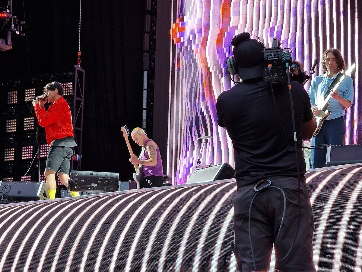 A stadium stage with a striped screen; a singer in red, a bassist and a guitarist