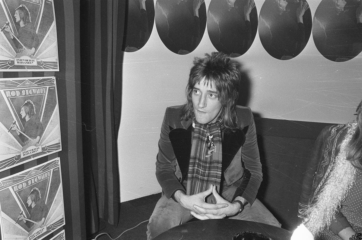 Rod Stewart in a velvet jacket and a tartan scarf, sitting with his hands together in front of framed gold records