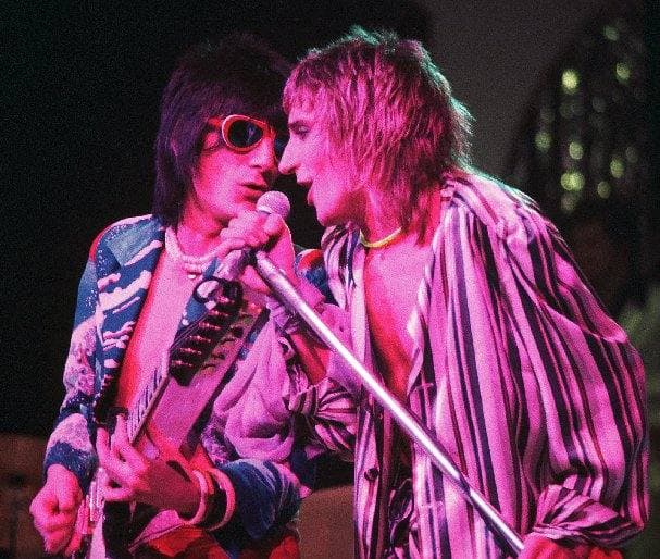 Ronnie Wood in sunglasses playing guitar and Rod Stewart in a striped shirt singing into one microphone, lit pink