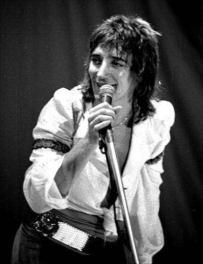 Rod Stewart in a white blouse and a belt of studs singing into a microphone on a stand