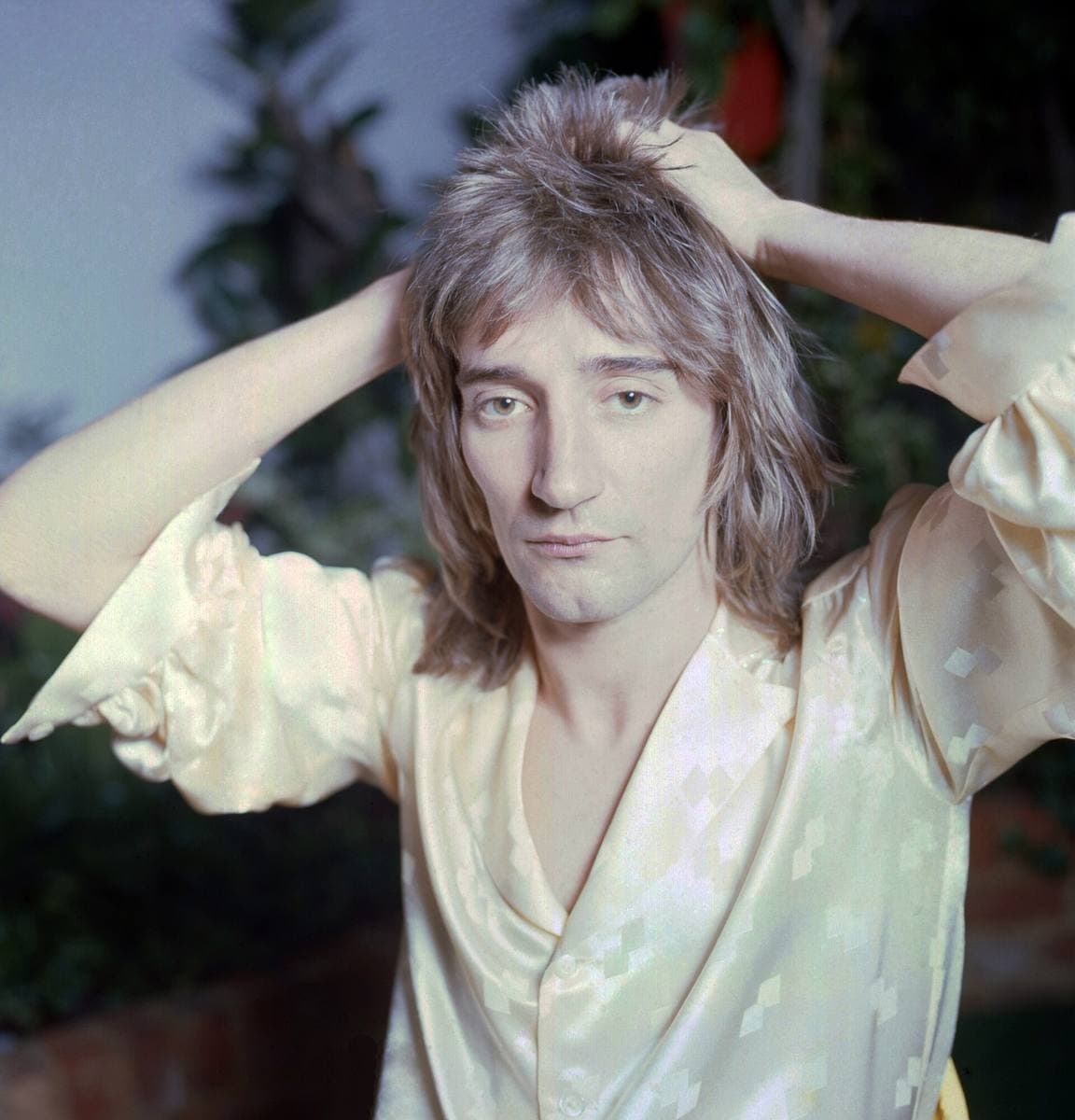 Rod Stewart with both hands pushed into his feathered hair, in a pale silk shirt, looking at the camera