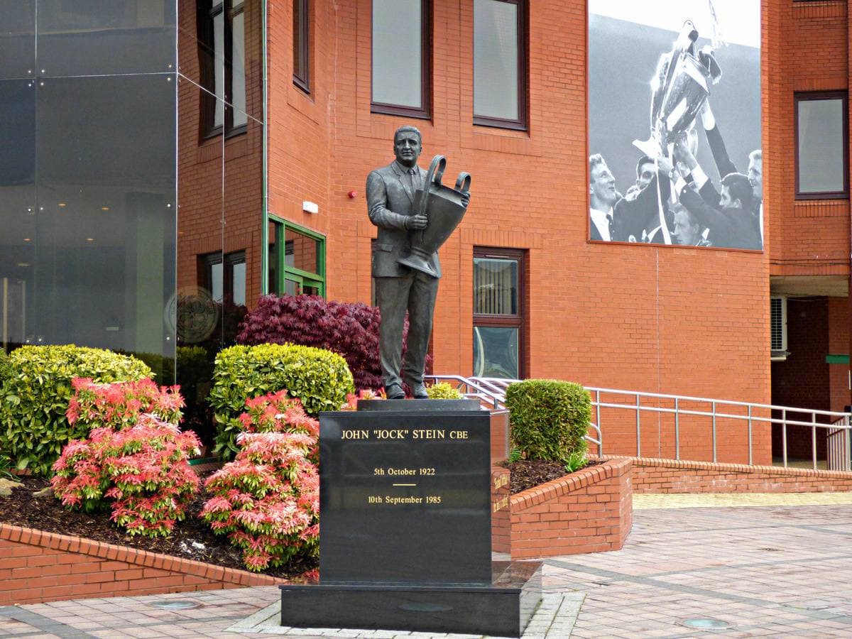 A bronze statue of a man holding a trophy on a dark plinth outside a red-brick stand