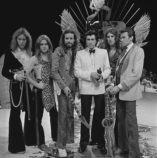 Six members of Roxy Music standing in a row in a television studio, Bryan Ferry in a white dinner jacket