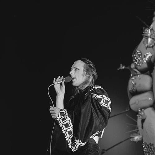 Brian Eno singing into a microphone in a patterned shirt against a black studio