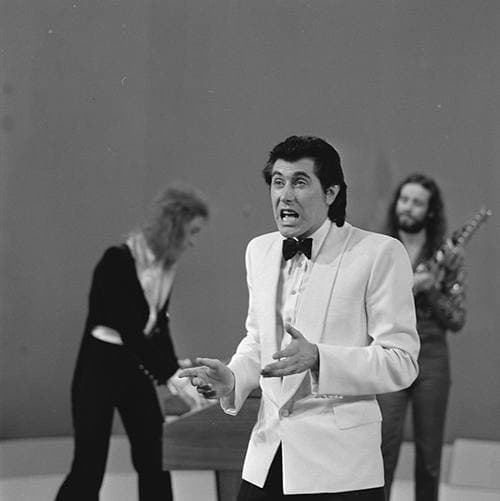 Bryan Ferry in a white dinner jacket and bow tie, singing with one hand out