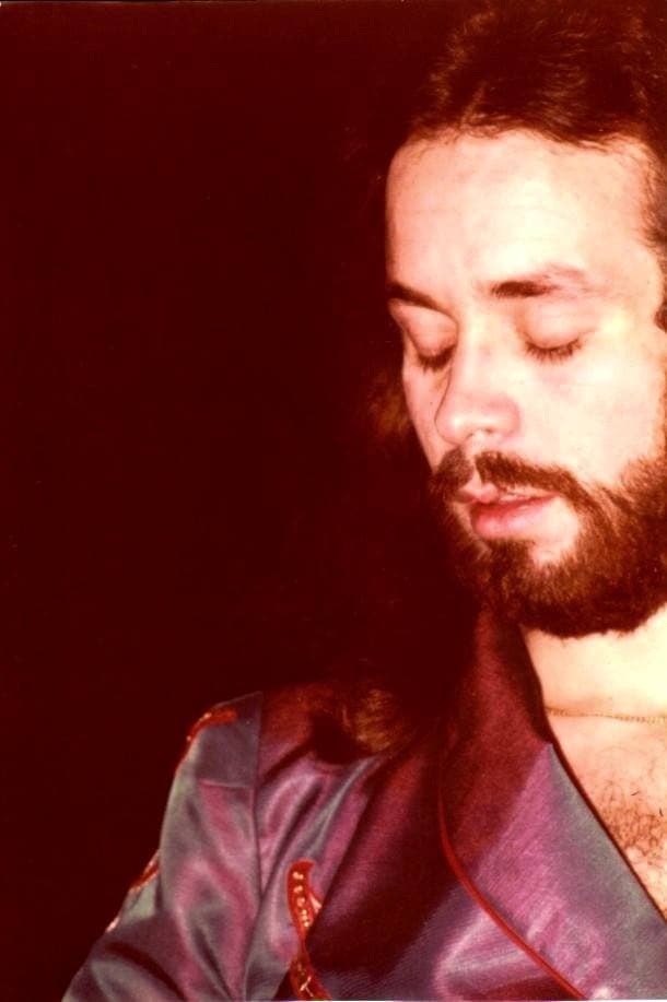 Phil Manzanera, bearded, looking down at his guitar under red stage light