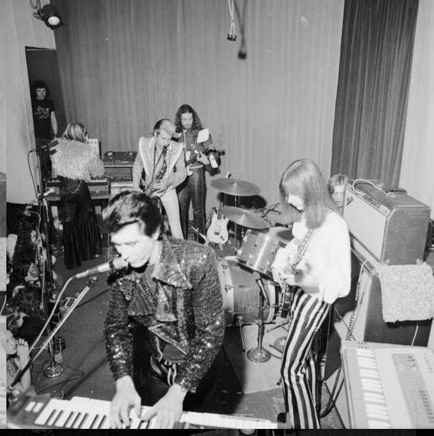 Bryan Ferry in a glittering jacket playing a keyboard on a small club stage, the band behind him