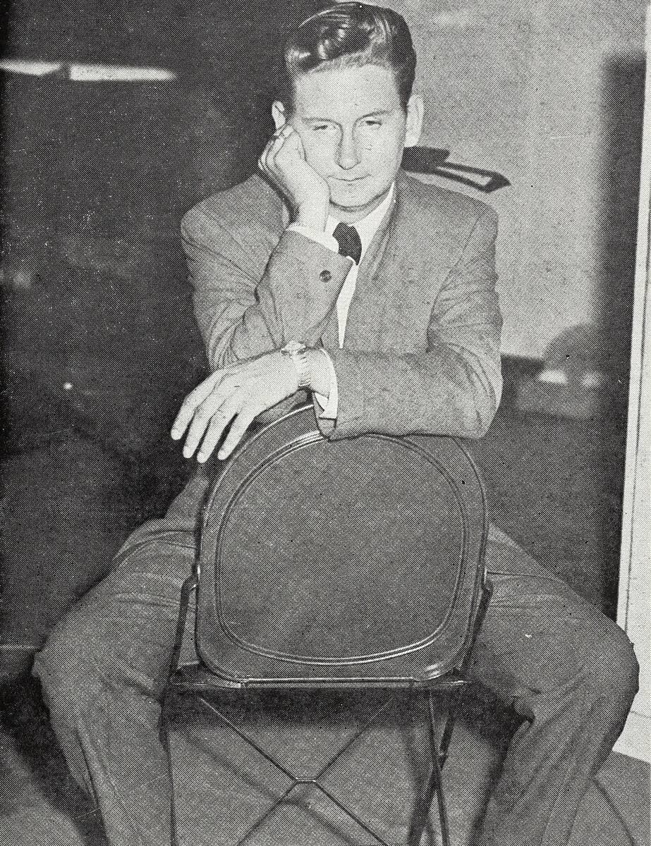 A young Roy Orbison in a suit and clear-rimmed glasses sitting astride a chair
