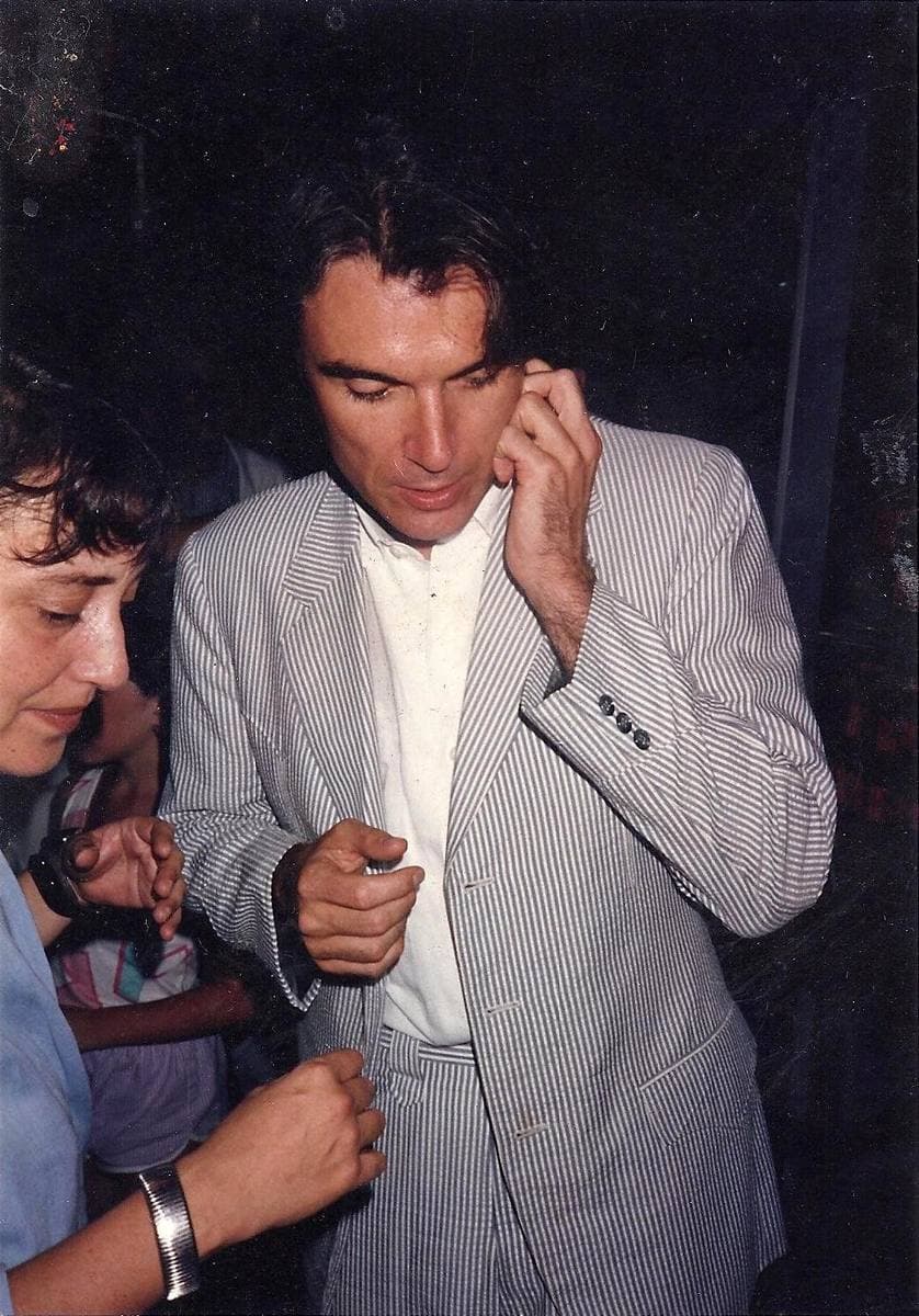 David Byrne in a pale striped jacket, talking with a woman in a crowded club