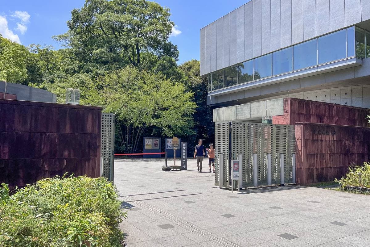 A paved entrance between brick-red walls and a modern glass building, with trees behind