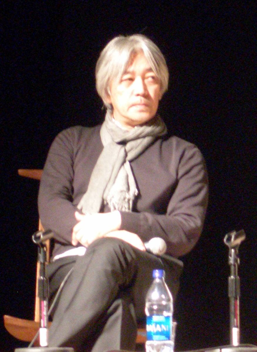 Ryuichi Sakamoto, grey-haired, in a dark jumper and scarf, seated on a stage with a microphone stand