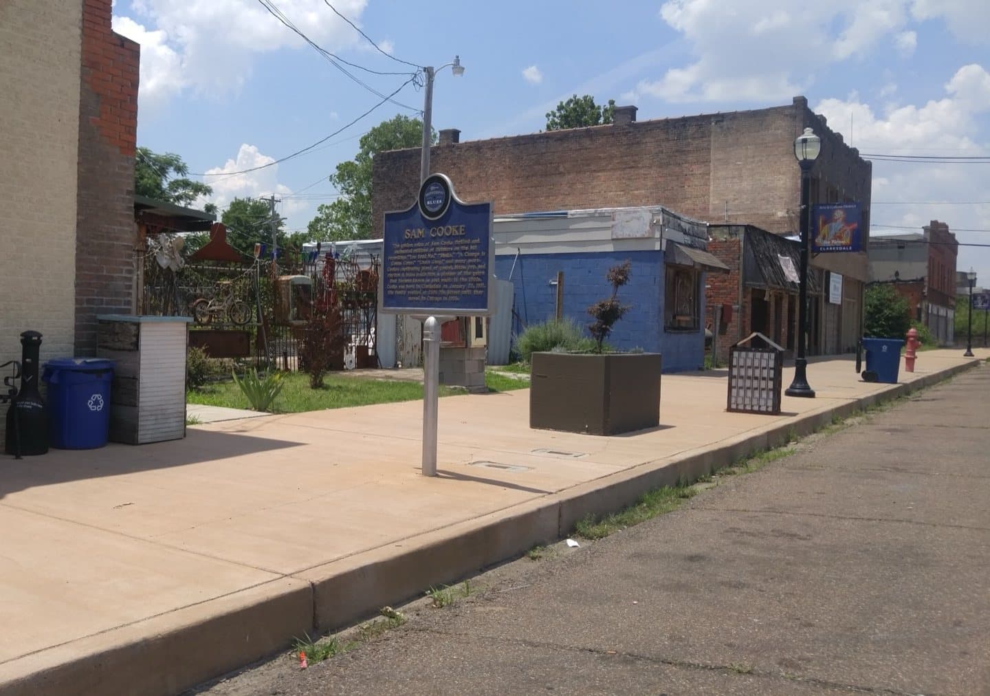A blue Mississippi Blues Trail marker on a pavement in downtown Clarksdale, beside a bench and low brick buildings