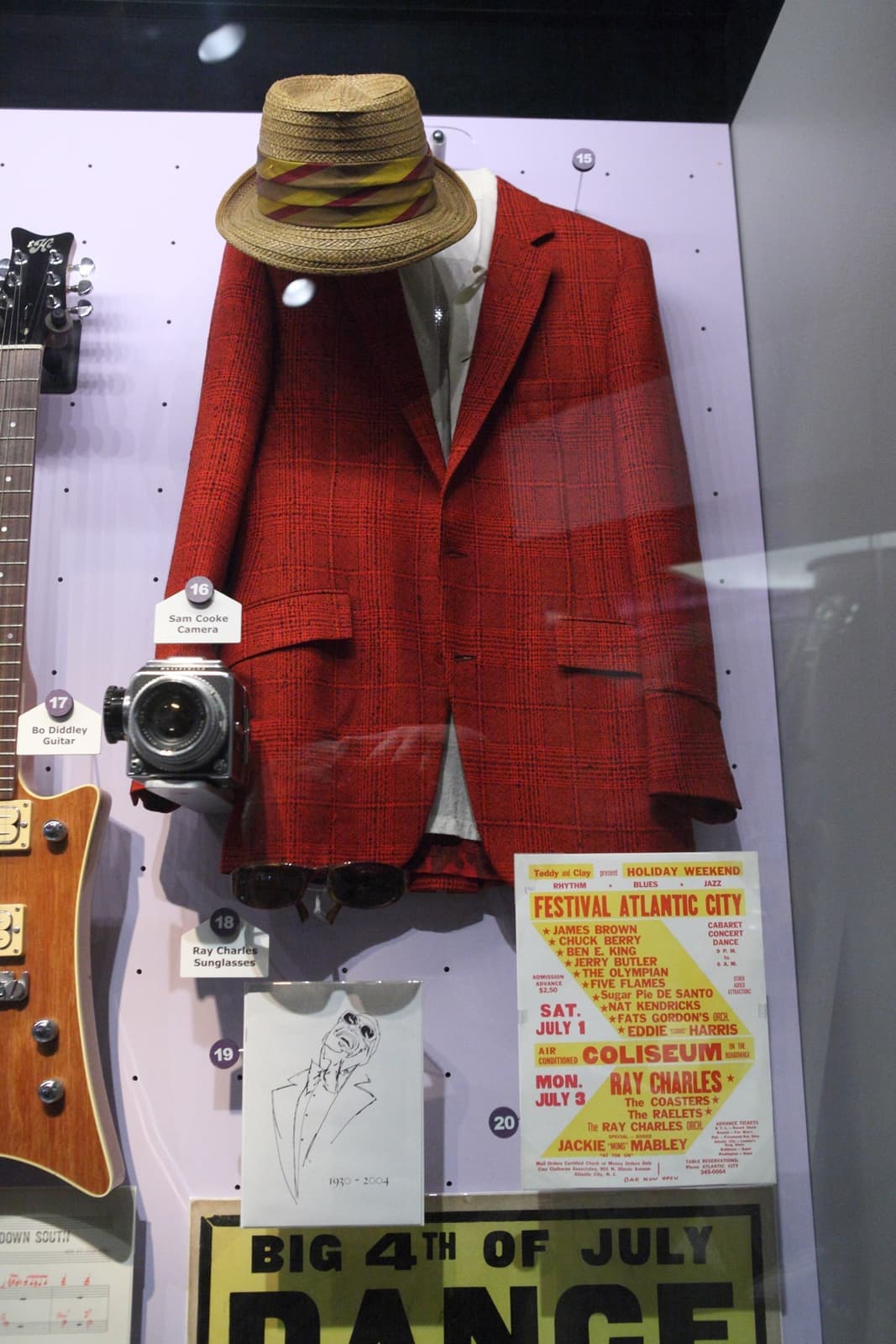 A red check jacket and straw hat displayed in a glass case with a camera and old concert posters