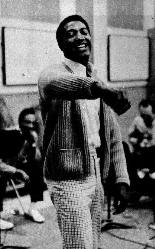 Sam Cooke laughing in a cardigan, one arm stretched out, with musicians seated around him in a studio