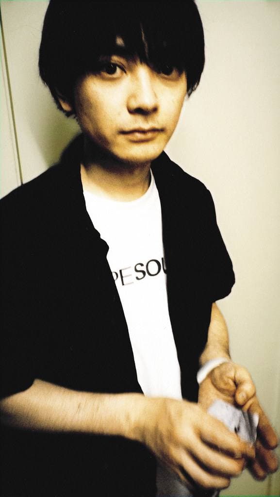 Keigo Oyamada sitting against a wall backstage, in a black shirt over a white T-shirt, looking at the camera