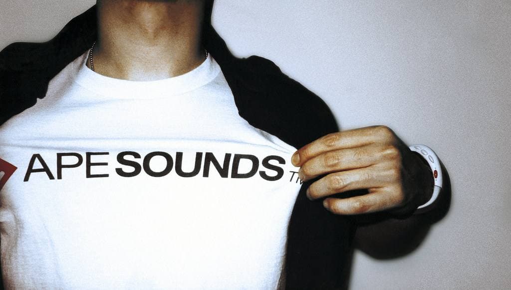 A man opening a dark jacket to show a white T-shirt printed Ape Sounds, a festival wristband on his wrist