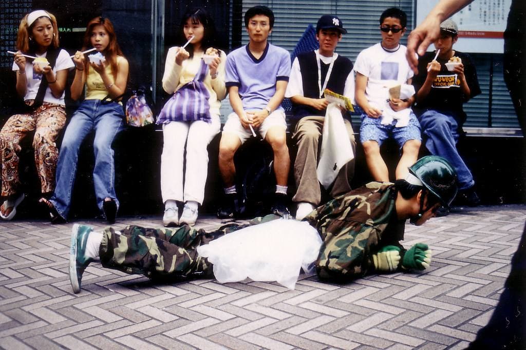 Teenagers in 1990s street clothes sitting in a row against a shop front while a young man in camouflage crawls along the paving