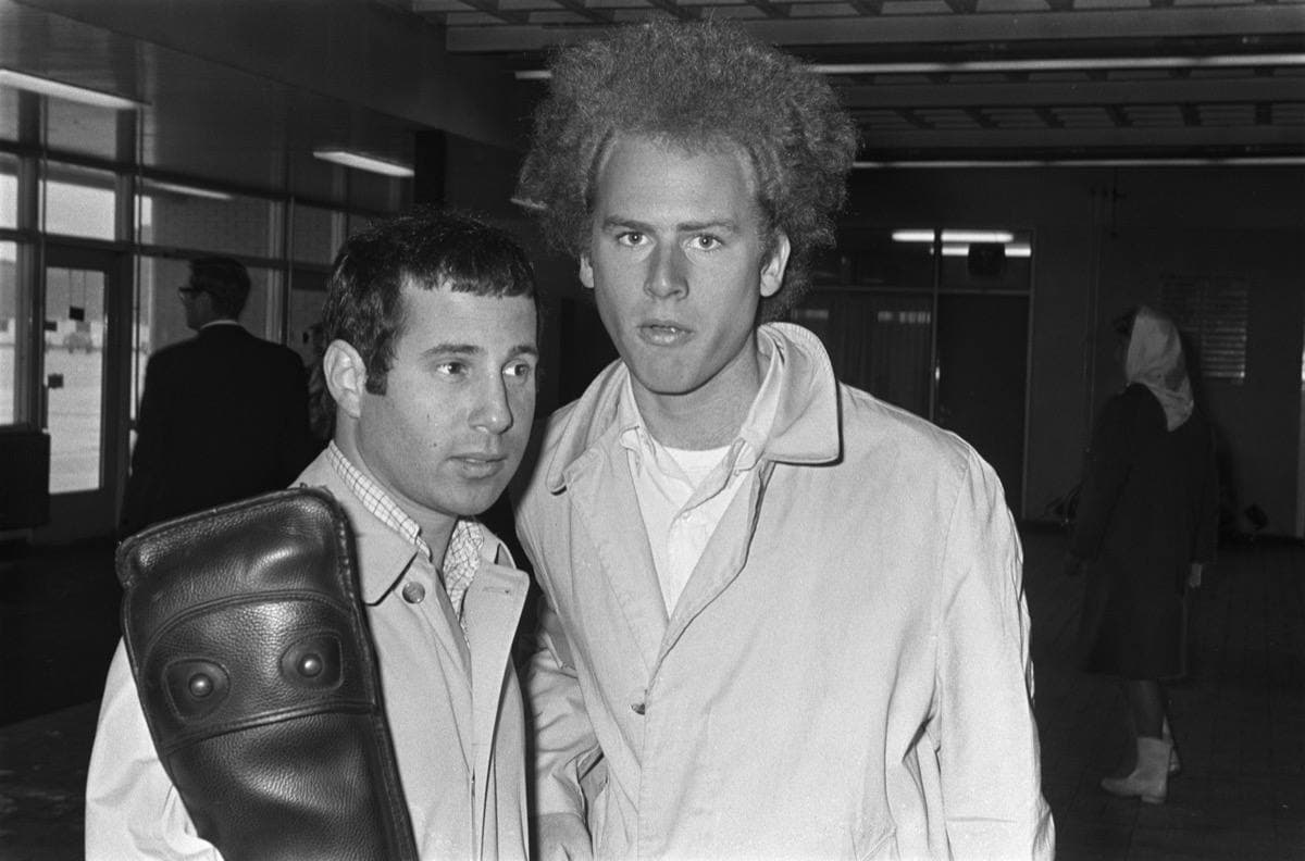 Paul Simon with a guitar case and Art Garfunkel in a raincoat in an airport hall