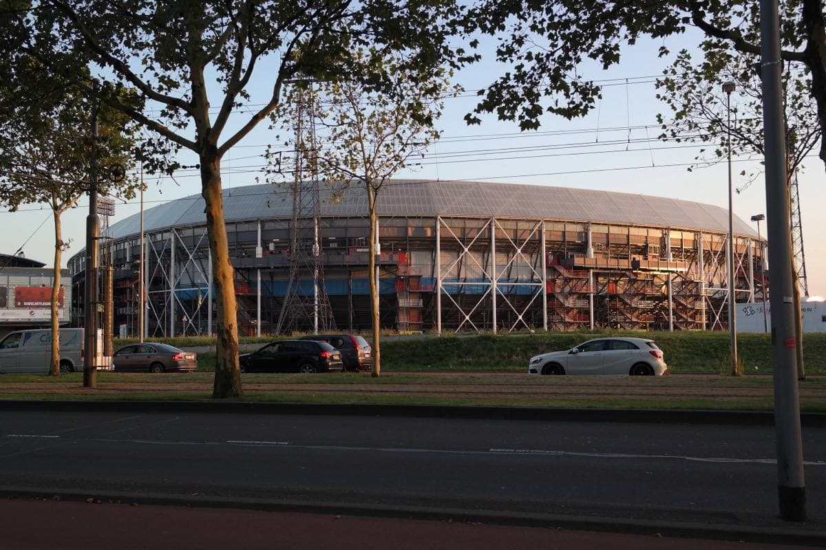 A round stadium with a steel exterior behind trees and a road