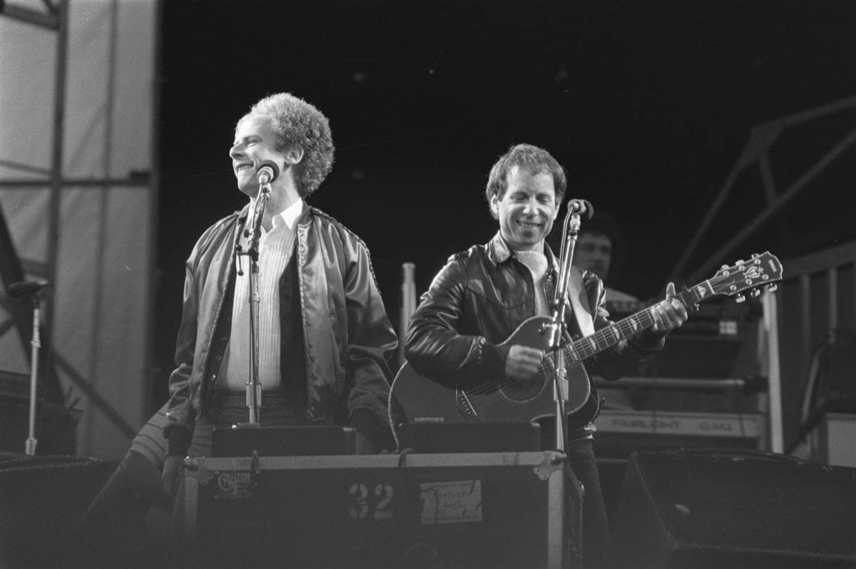 Art Garfunkel and Paul Simon, with a guitar, laughing at their microphones on an outdoor stage