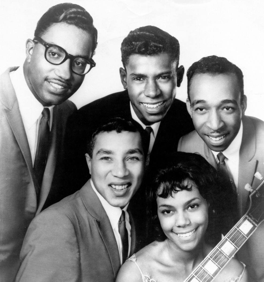Five young people in a tight publicity portrait: four men in suits and a smiling woman at the front with a guitar neck beside her
