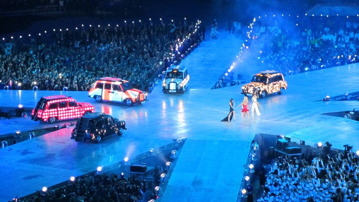 London taxis covered in lights driving across a lit stage in a packed stadium