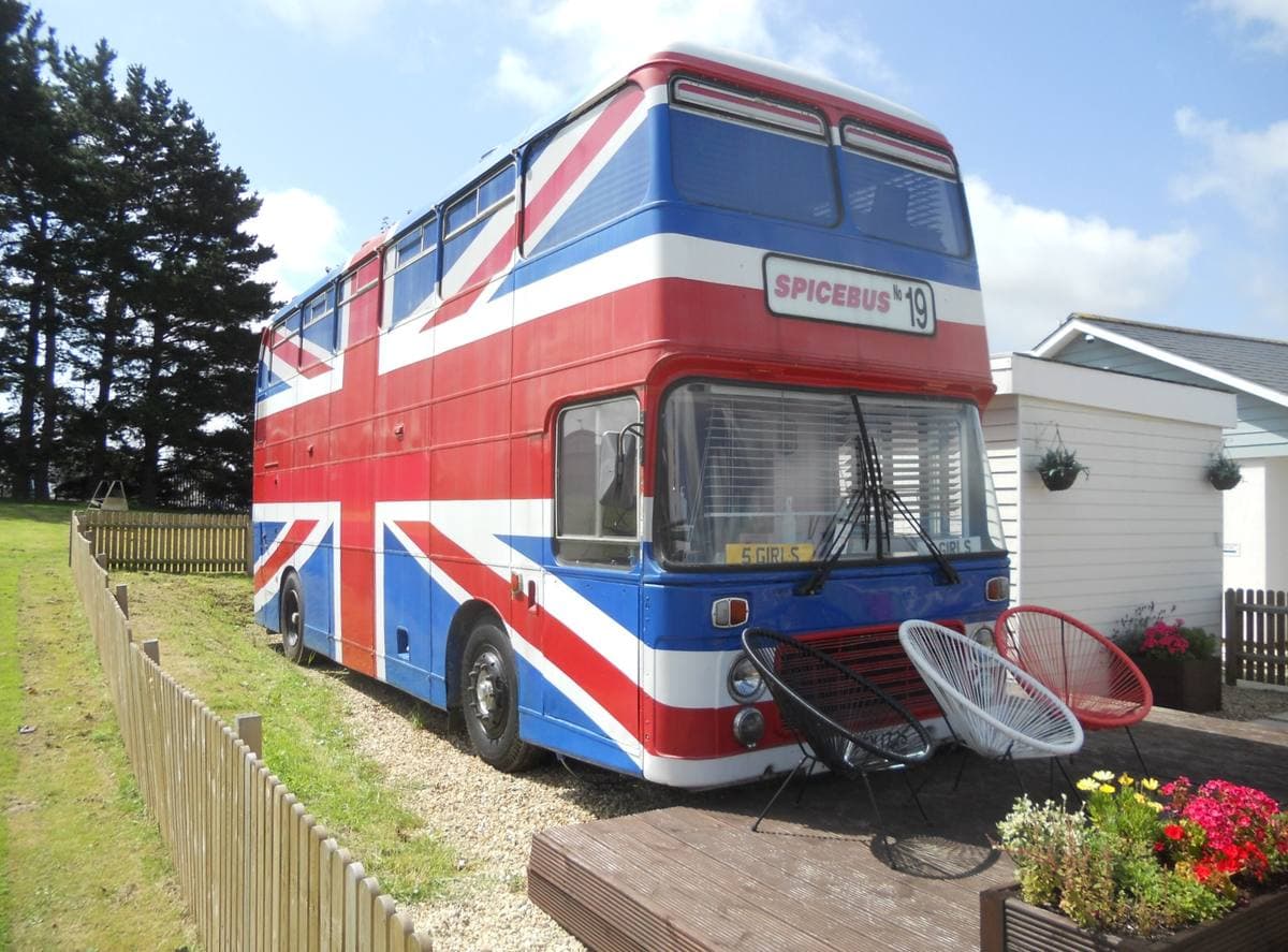 A double-decker bus painted red, white and blue parked on gravel beside a fence