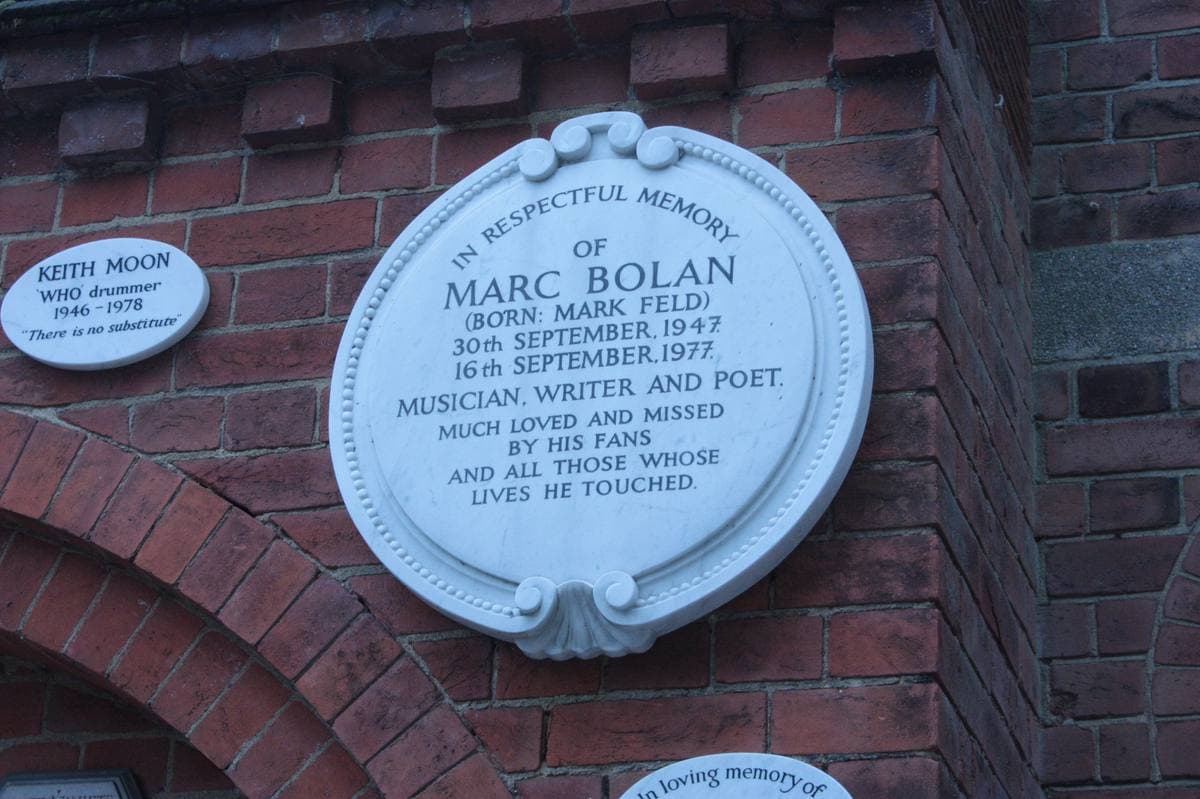 A white oval memorial plaque to Marc Bolan on a red brick wall