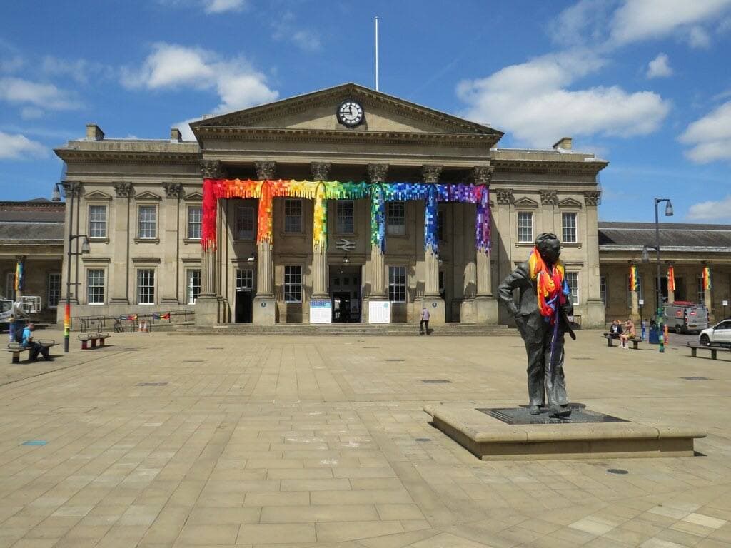 A wide stone square in sunshine with the columned railway station behind and a statue in front