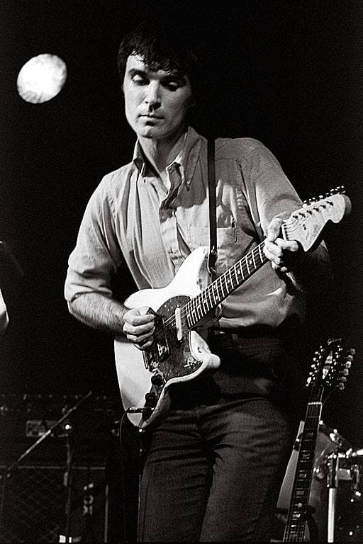 David Byrne playing an electric guitar on a small stage, black and white